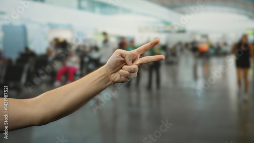 Man gesturing peace sign indoors at busy airport with flight passengers in background, showcasing travel atmosphere and human connection.