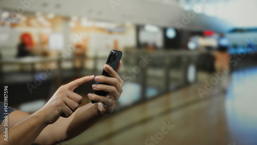 Obraz na płótnie Man holding smartphone in indoor shopping mall with blurred background, focusing on touchscreen, reflecting modern retail technology and communication. w salonie