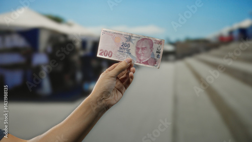 Hand holding turkish lira banknote on a sunny day in an outdoor market, highlighting commerce and shopping on busy street.