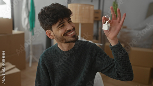 Obraz na płótnie Young man smiling in new home surrounded by boxes holding house key in living room indicating successful moving and new beginnings w salonie