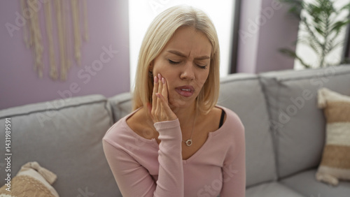 Obraz na płótnie Woman holding cheek in pain sitting on a couch in a cozy living room with purple walls, showcasing a stylish and modern home interior with natural light. w salonie