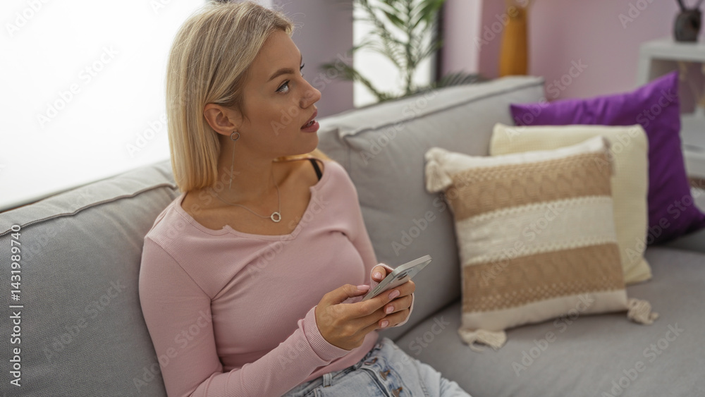 Young woman sitting on comfortable sofa in bright living room holding smartphone with curious expression surrounded by pillows in stylish home interior