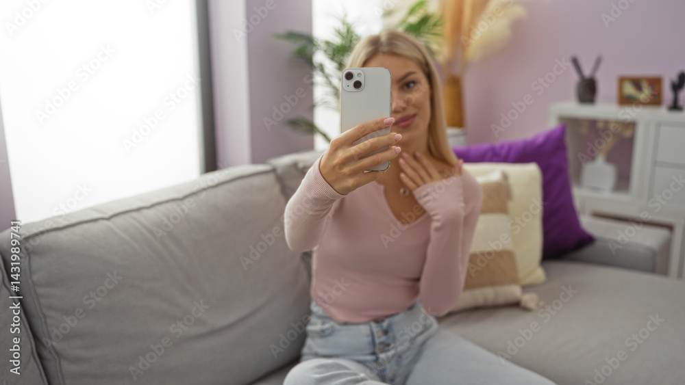 Woman taking selfie indoors, sitting in stylish living room, blonde hair, wearing casual pink shirt, smiling, relaxed home atmosphere, modern apartment decor, young caucasian adult