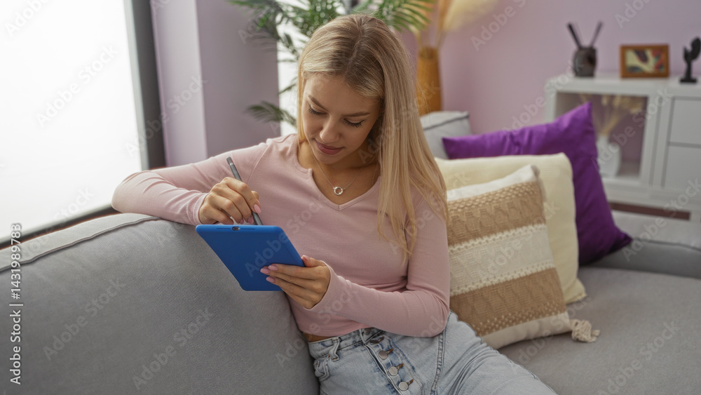 Woman writing on tablet in cozy living room, conveying modern indoor lifestyle; young and blonde, relaxing at home, focused on screen artistry, embracing technology comfortably.