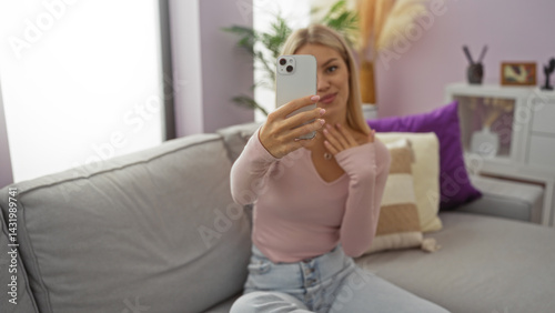 Obraz na płótnie Woman taking selfie indoors, sitting in stylish living room, blonde hair, wearing casual pink shirt, smiling, relaxed home atmosphere, modern apartment decor, young caucasian adult w salonie
