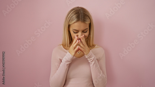Woman contemplating blonde hair pink sweater against soft pink background, expressing serene introspection with hands together showing thoughtful gesture.