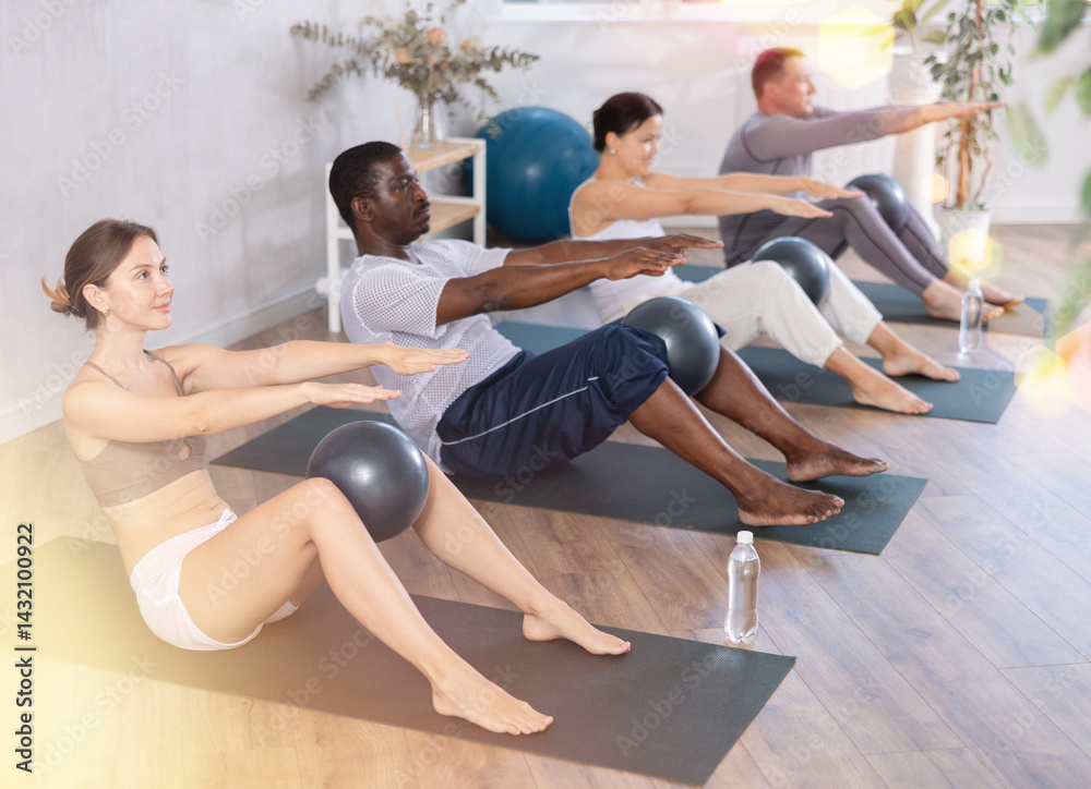 Obraz premium Woman in activewear sitting on yoga mat during group workout with fitness ball at modern gym. Healthy lifestyle concept