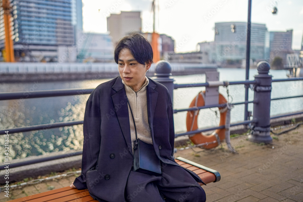 A Japanese man in his twenties wearing a black jacket is in Minato Mirai, Yokohama, Kanagawa Prefecture. He sits on a bench using his smartphone, with a backdrop of high-rise buildings around him.