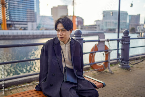 Obraz na płótnie A Japanese man in his twenties wearing a black jacket is in Minato Mirai, Yokohama, Kanagawa Prefecture. He sits on a bench using his smartphone, with a backdrop of high-rise buildings around him. w salonie