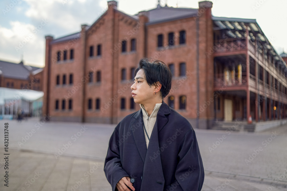 A Japanese man in his twenties stands in Minato Mirai, Yokohama, Kanagawa Prefecture, looking ahead with a red brick building by the sea behind him, where past and present gracefully come together.