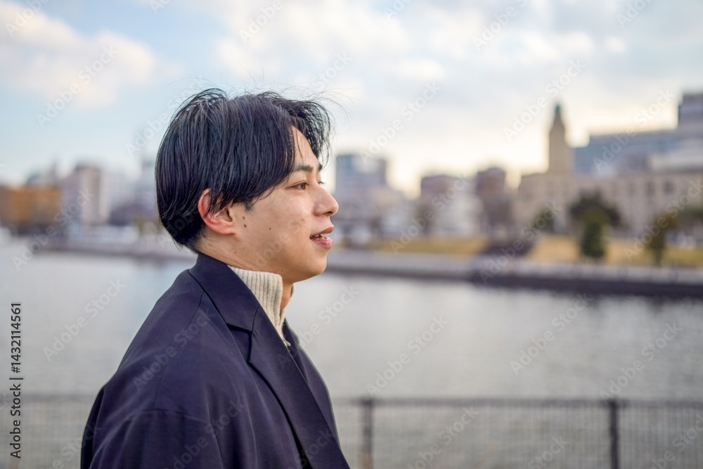 A Japanese man in his twenties wearing a black jacket walks along a seaside path in Minato Mirai, Yokohama, Kanagawa Prefecture, enjoying the breeze, city buildings, and the calm harbor atmosphere.