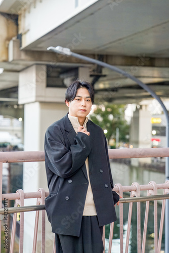 Obraz na płótnie A Japanese man in his twenties wearing a black jacket is in Minato Mirai, Yokohama, Kanagawa Prefecture. He sits on a bench, gazing into the distance, surrounded by the calm mood of the modern city. w salonie