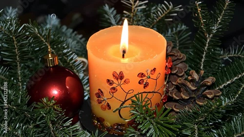 Softly glowing orange candle decorated with a floral pattern stands among fresh green pine branches