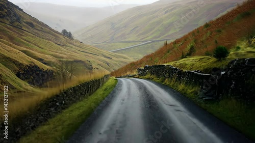Winding asphalt road cuts through a lush green valley, flanked by hills and a stone wall, under an overcast sky