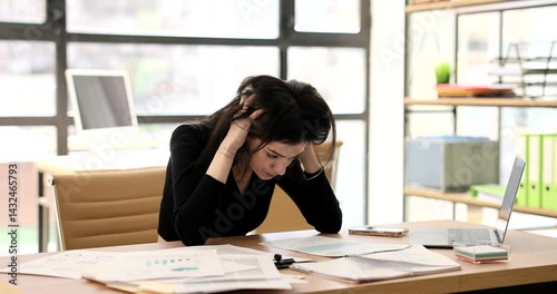 Woman sits at desk in office looking stressed and overwhelmed. Brunette employee with hands on head and papers on desk suggesting work-related stress