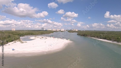 Drone video moves backwards over Wiggins Pass a beach river opening with boats moored on the beach. Trees surround and a residential community can be seen inland. Sunny, bright day with clouds above.