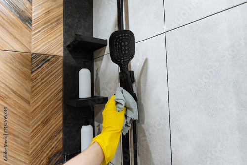 In a brightly lit bathroom, an individual wearing yellow rubber gloves diligently scrubs the shower head to ensure that it is clean, shiny, and functioning properly after heavy usage
