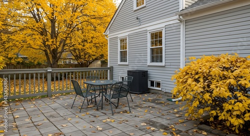 Autumn patio scene with a gray house in the background.  A table and chairs sit on a stone patio, surrounded by fall foliage.