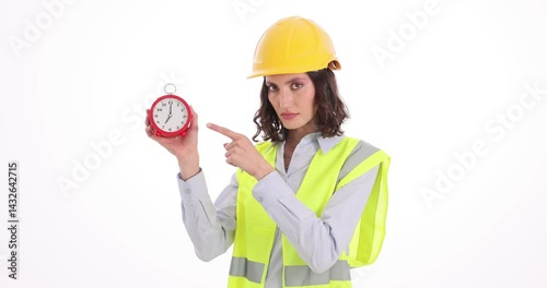 Forewoman in helmet shows alarm clock threatening by finger on white background. Female taskmaster reminds about deadline at construction site