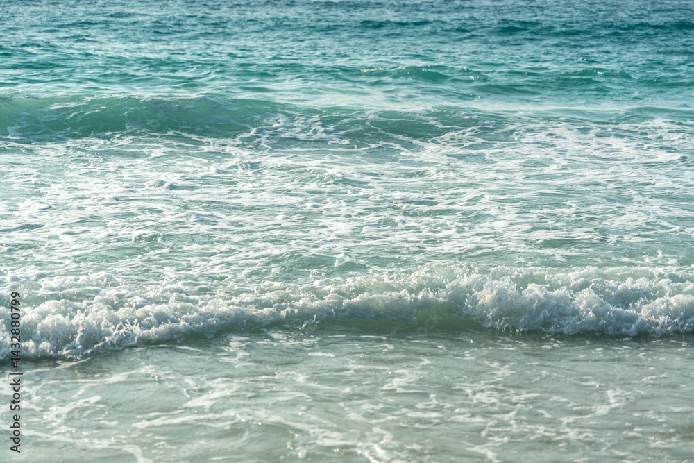 Close-up texture of ocean waves and sandy beach under sunlight with turquoise tones and soft foam
