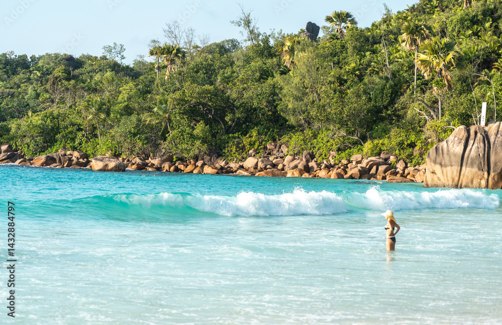 Woman in bikini and hat stands in turquoise sea at a tropical Seychelles beach with granite boulders