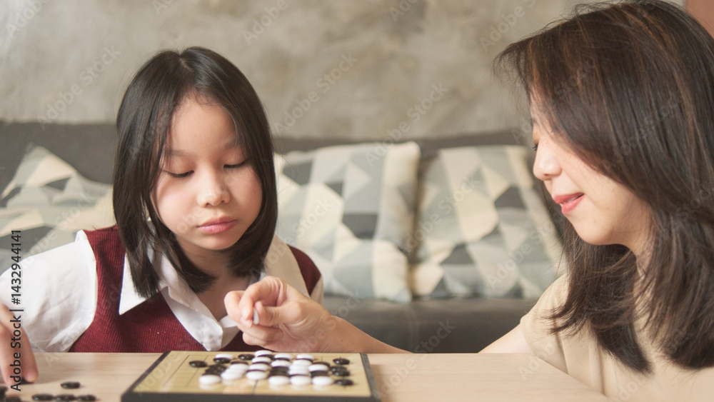 Fototapeta premium Asian family, mum, and kid fun playing Go, checkers board game on table in home living room together with love and cheerful, happy weekend relaxing hobby of child and parent, concentrate and compete.
