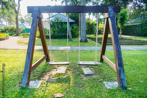 Two empty wooden swings hang in a tranquil park setting with green grass and surrounding trees on a sunny day