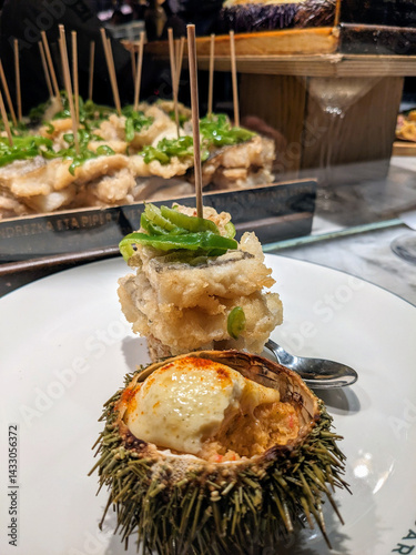 Delectable seafood tapas features fried fish topped with green peppers alongside a sea urchin on a plate.