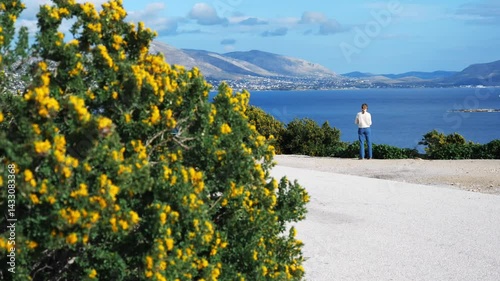 Bright yellow flowers bloom in the foreground as a woman in a white sweater and jeans stands on a viewpoint, enjoying the breathtaking sea view. The vibrant blue sky and sea contrast with the flowers