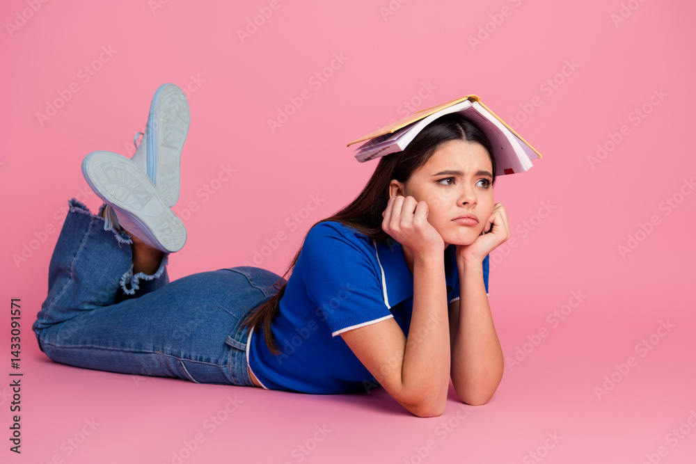 Young female student lying on pink background with book on head, showing expressions of frustration