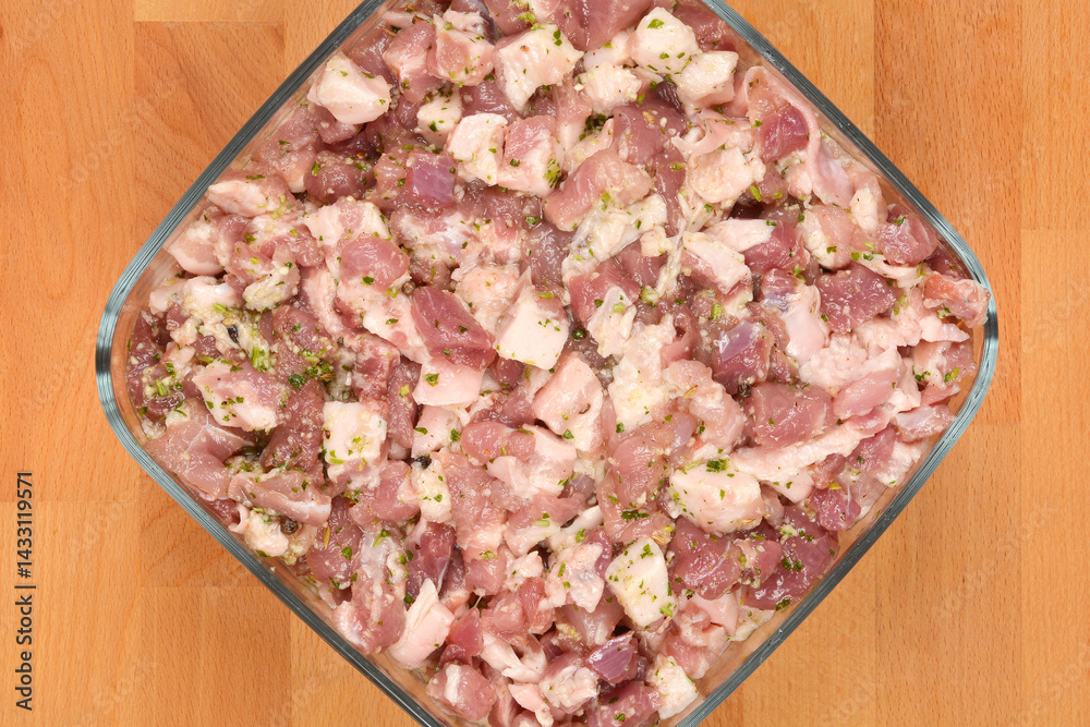 Diced and seasoned pork meat, prepared with spices for sausage making, fills a square glass dish on a wooden surface
