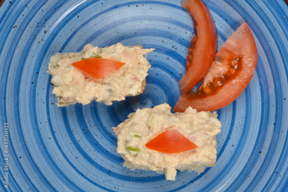 Two small bread slices topped with tuna, cream cheese, egg, caper, onion, and mustard spread are garnished with tomato on a blue plate