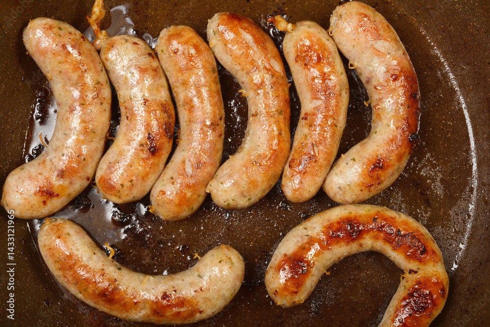 Several plump homemade sausages are sizzling in a dark frying pan, browning nicely in their own rendered fat