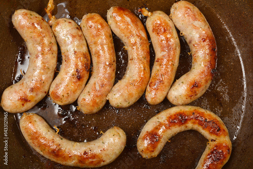 Several plump homemade sausages are sizzling in a dark frying pan, browning nicely in their own rendered fat