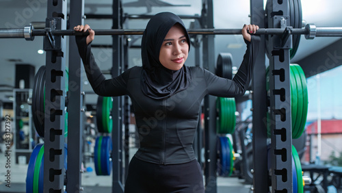 A confident Southeast Asian Muslim woman in a black sports hijab preparing to lift a barbell in a modern gym. The image highlights strength, empowerment, and diversity in fitness culture.