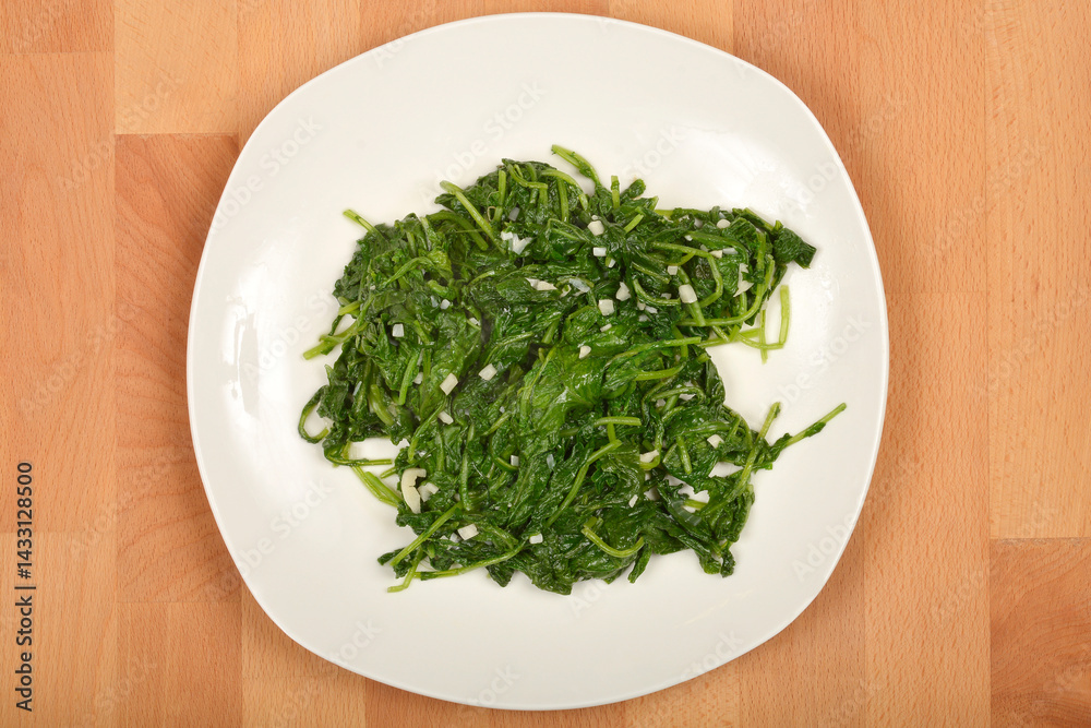 A serving of cooked radish leaves with pieces of garlic is presented on a white plate atop a wooden surface, captured from above