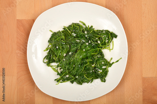 Obraz na płótnie A serving of cooked radish leaves with pieces of garlic is presented on a white plate atop a wooden surface, captured from above w salonie