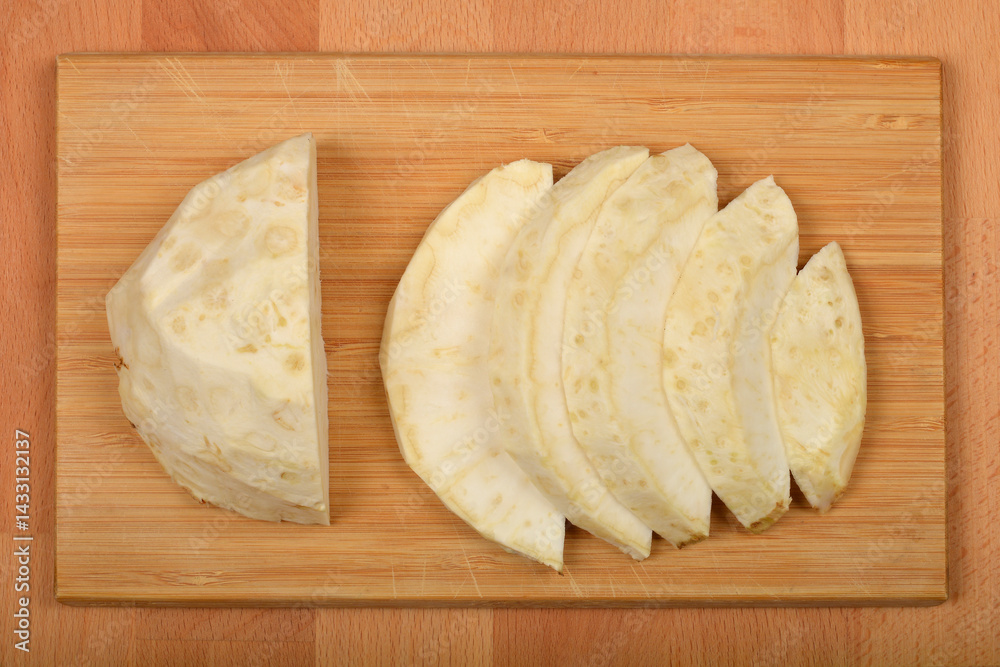 Slices and a wedge of raw, creamy white celeriac are arranged on a light brown wooden cutting board, ready for preparation
