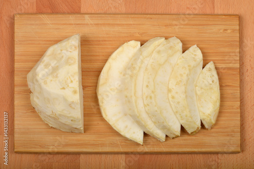 Slices and a wedge of raw, creamy white celeriac are arranged on a light brown wooden cutting board, ready for preparation