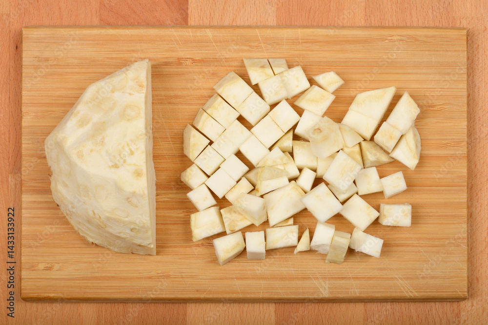 A peeled wedge and a pile of diced raw celeriac are presented on a light brown wooden cutting board, illustrating different cutting styles