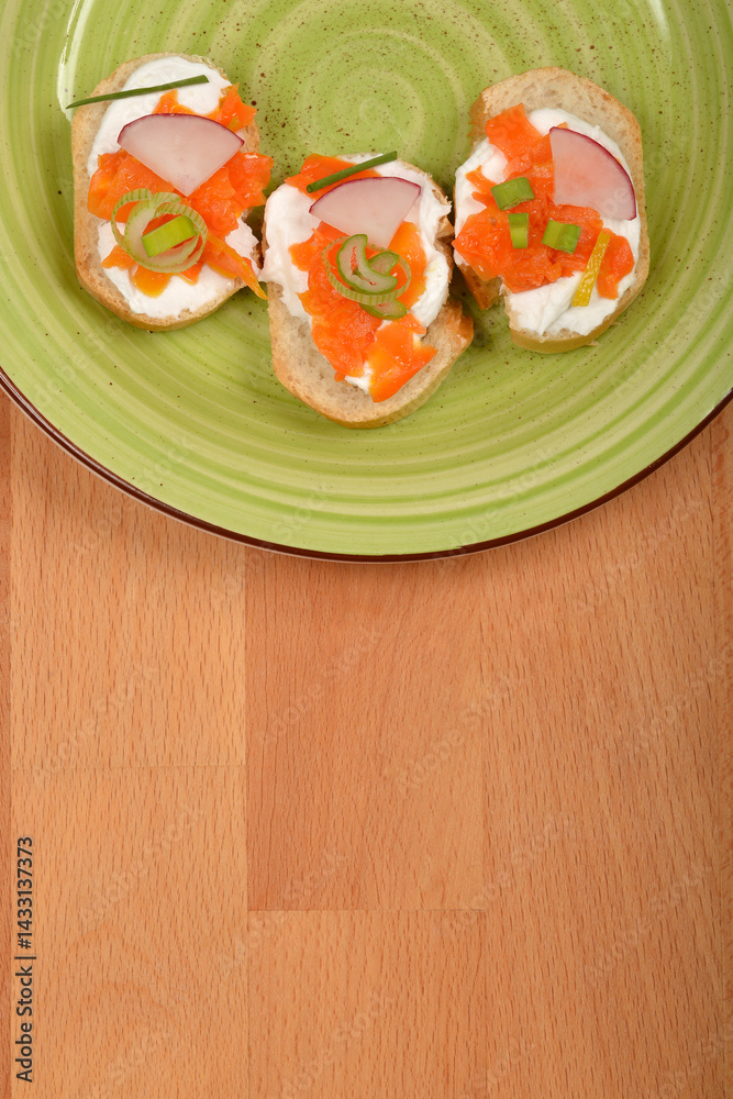 An overhead shot shows three elegant canapes with a creamy spread, vibrant orange carrot prepared as vegan salmon, radish slices, and green chives on a light green plate with a dark rim, set on a wood