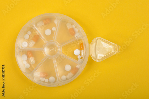 Obraz na płótnie A clear, round pill organizer with one compartment open and pills inside is positioned on a bright yellow surface w salonie