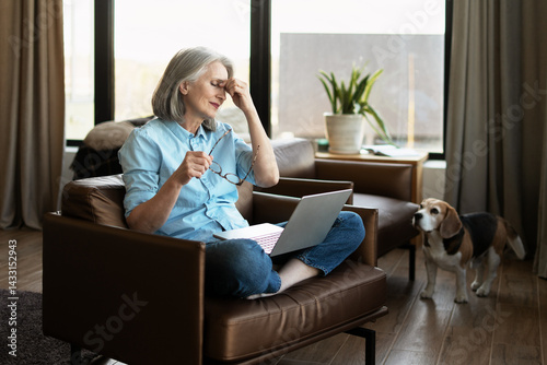 Stressed senior woman having headache while working from home with laptop photo