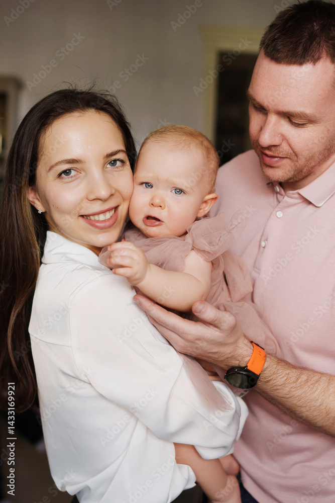 Happy and gentle parents hold baby daughter in pink dress in their arms.