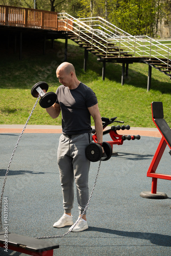 Man lifting dumbbells at an outdoor gym, focused on strength training. Wearing athletic clothing. Fitness equipment in outdoors park setting. Urban sports ground