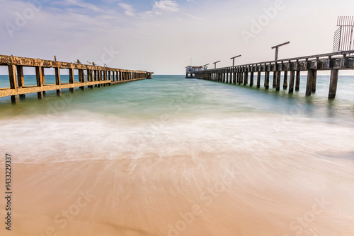 Rustic parallel piers stretch out into the ocean with gentle waves washing onto the sandy beach under a partly cloudy sky