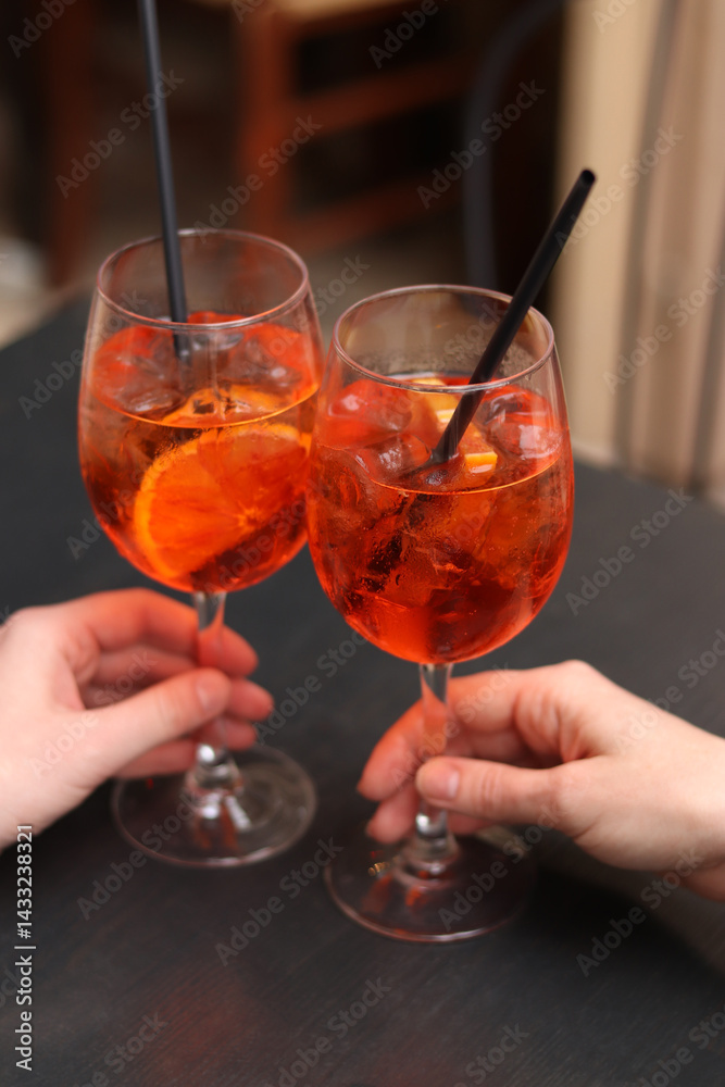 Two glasses with Aperol Spritz in the hands of girls. Cold refreshing alcoholic cocktail Aperol Spritz. Cocktails close-up with selective focus. Alcoholic drink with a straw. Traditional Italian drink
