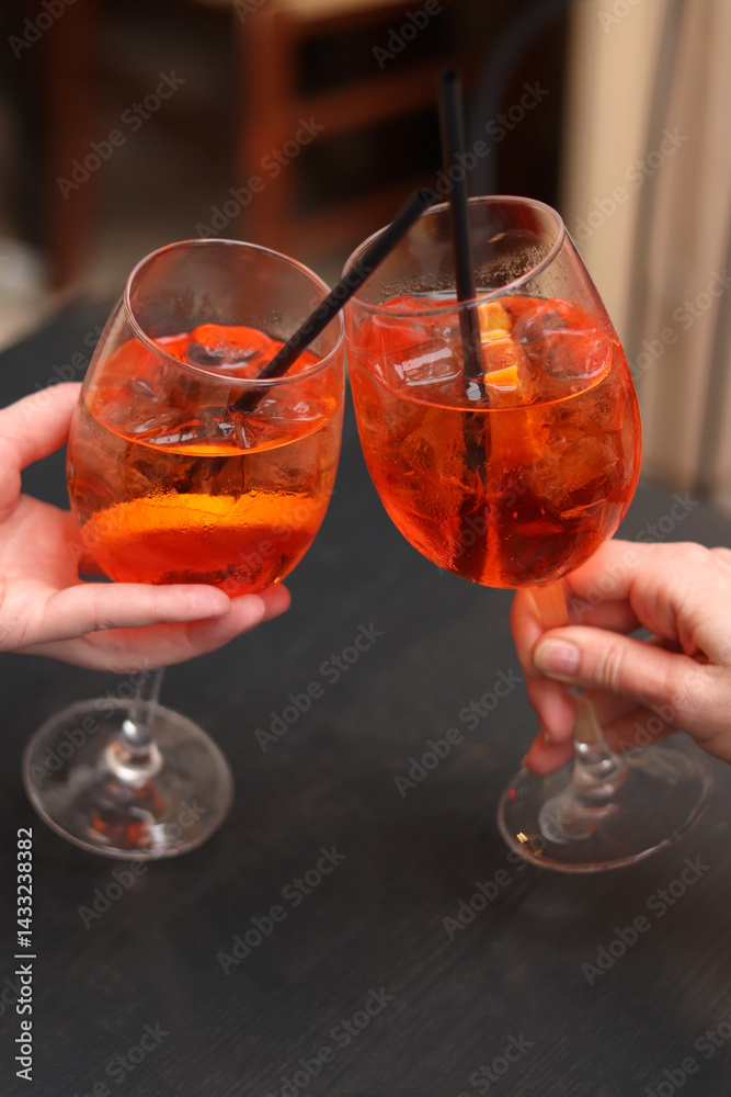 Two glasses with Aperol Spritz in the hands of girls. Cold refreshing alcoholic cocktail Aperol Spritz. Cocktails close-up with selective focus. Alcoholic drink with a straw. Traditional Italian drink
