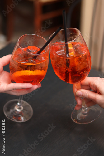 Obraz na płótnie Two glasses with Aperol Spritz in the hands of girls. Cold refreshing alcoholic cocktail Aperol Spritz. Cocktails close-up with selective focus. Alcoholic drink with a straw. Traditional Italian drink w salonie