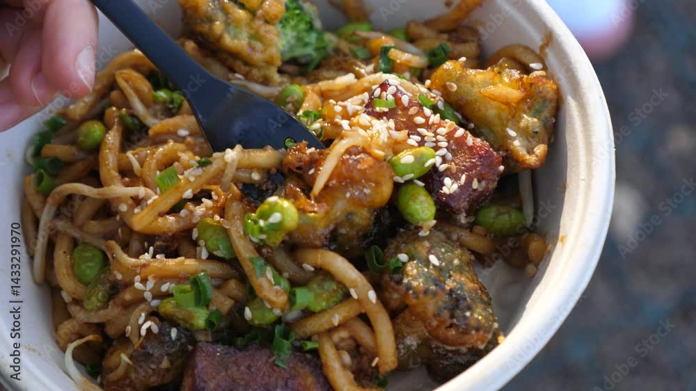Disposable bowl of udon noodles with tempura broccoli, edamame, green onions, sesame seeds and teriyaki sauce being enjoyed with a black plastic fork